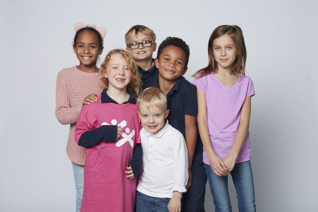Six diverse children of various ages pose together and smile for a studio portrait against a plain white background.