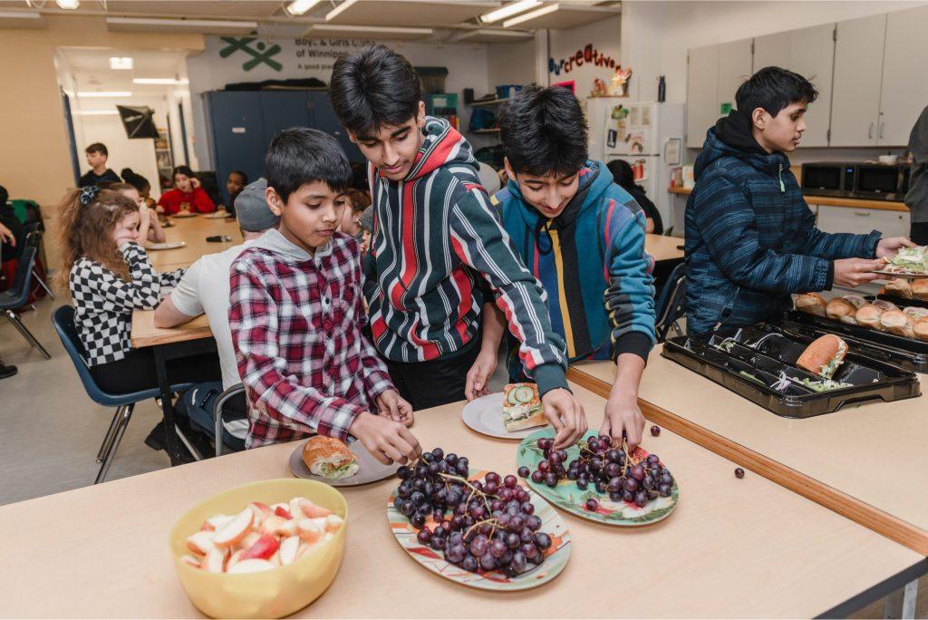 Four boys gather around a cafeteria table, with three of them selecting sandwiches and red grapes from platters while a fourth boy prepares a tray of subs in the background.