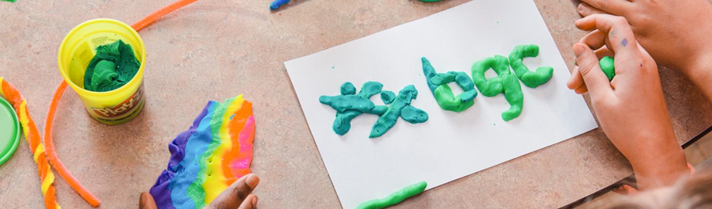 banner-placeholder Top-down view of hands using green modeling clay to spell out "#bgc" on a white piece of paper next to a piece of rainbow-colored clay and craft supplies.