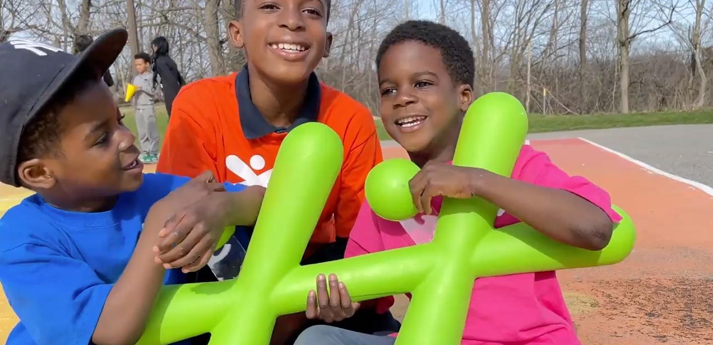 Three young boys laugh together outdoors while holding a large, bright green inflatable version of the BGC logo.