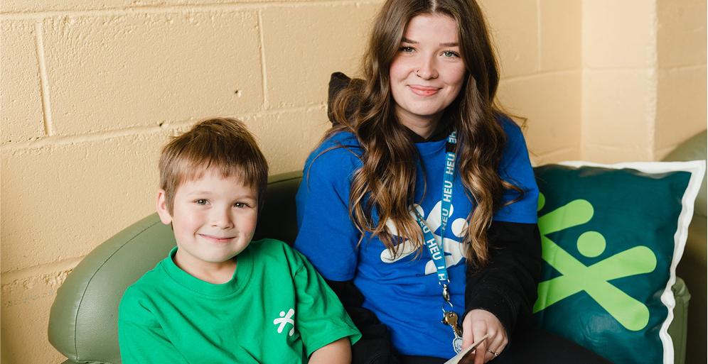 A young woman with long wavy brown hair and a boy in a green t-shirt sit together on a green couch in front of a light yellow brick wall.