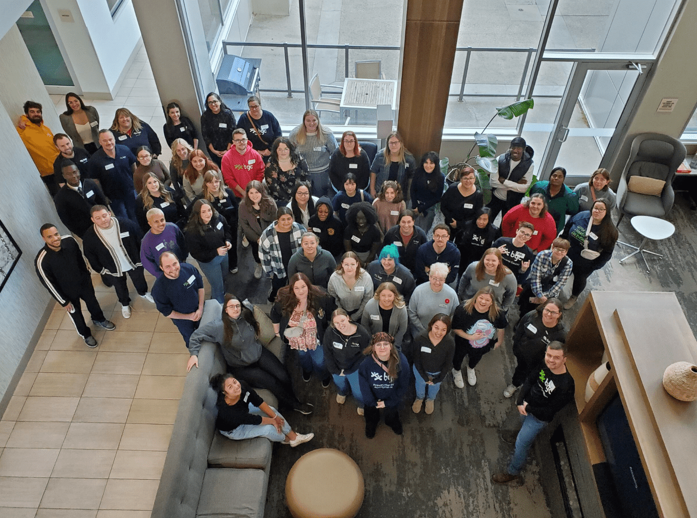 A large group of approximately fifty diverse people stand and sit together for a group photo in a modern, sunlit lobby featuring high ceilings and large windows.