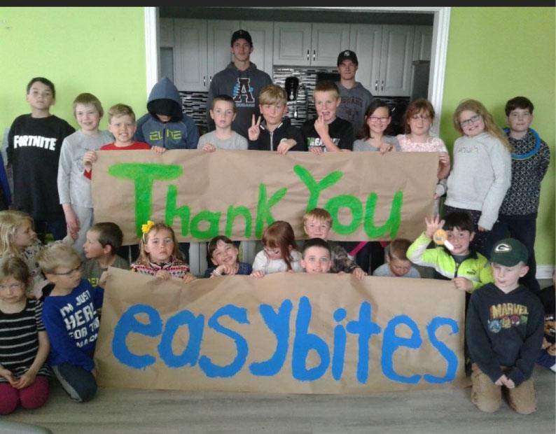 A large group of children pose together holding handmade brown banners that read "Thank You easybites" in green and blue paint.