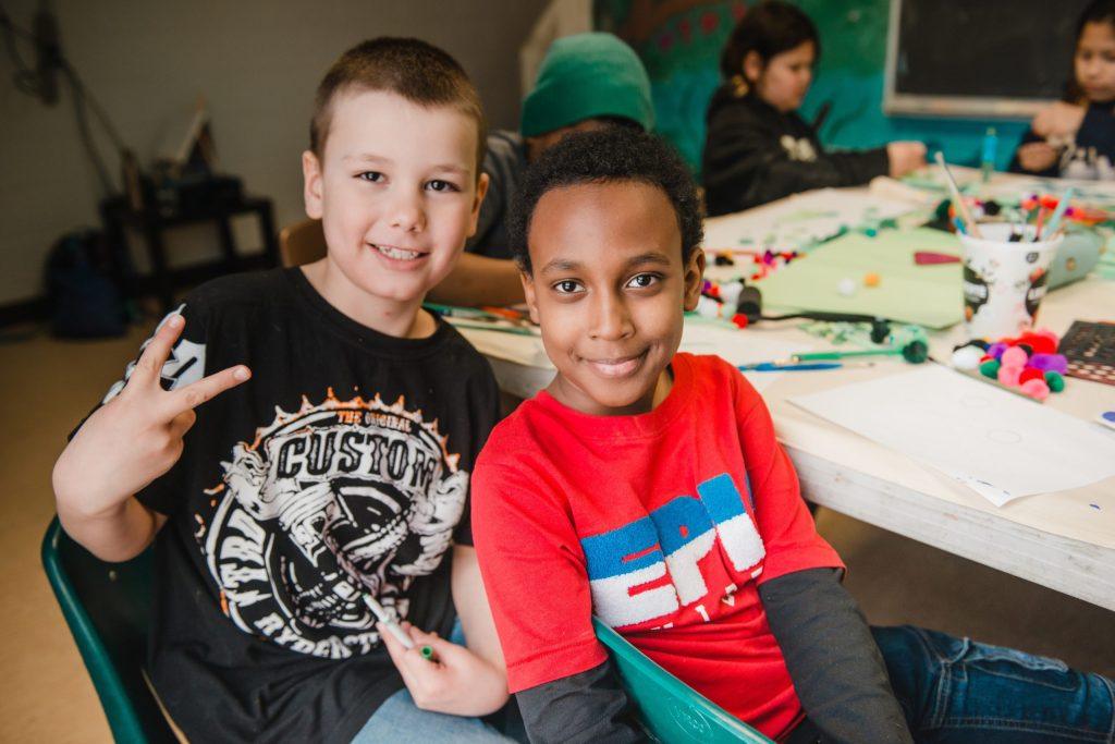 Two young boys sitting at a table with arts and crafts supplies, one making a peace sign.