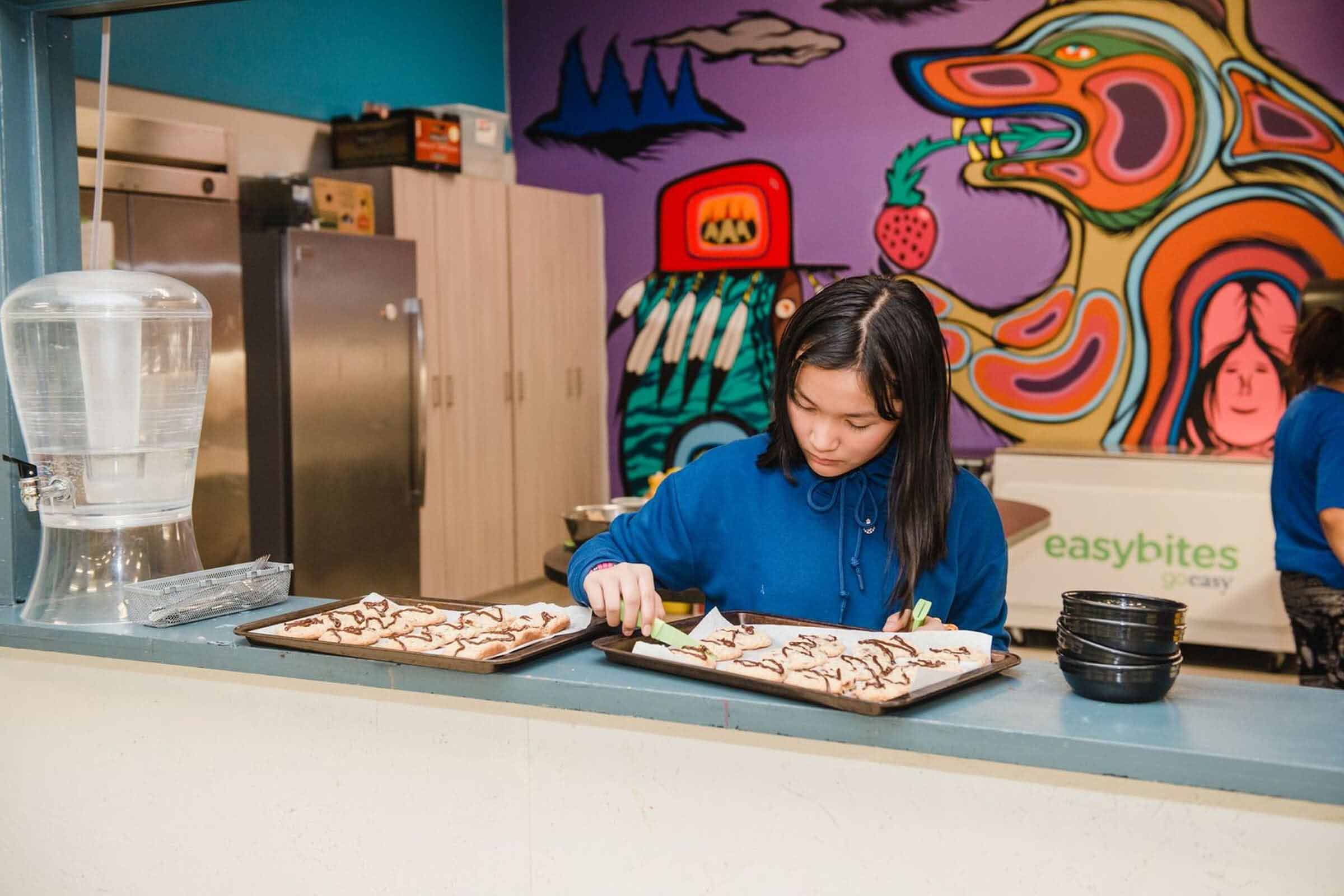 Young girl preparing baked snacks at a BGC kitchen with colourful mural in the background.