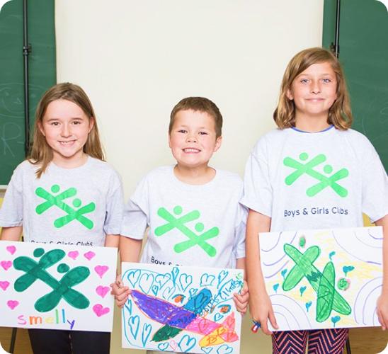 Three smiling children wearing grey "Boys & Girls Clubs" t-shirts proudly hold up their colorful, hand-drawn artwork.