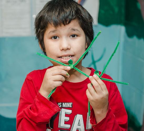 Young boy in a red shirt smiling while holding a green pipe cleaner craft in front of him.