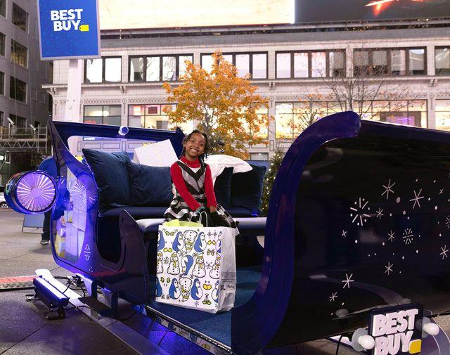 A young girl smiles while sitting in a festive sleigh display during Best Buy’s holiday activation event, holding a branded gift bag.
