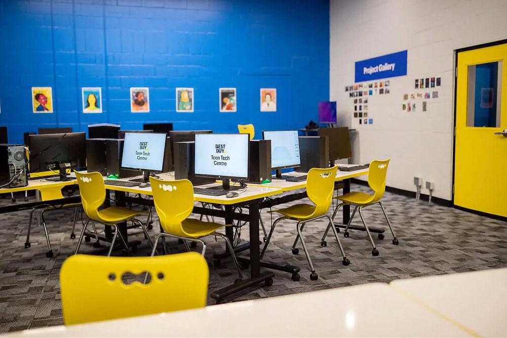 A modern computer lab at a Best Buy Teen Tech Centre featuring rows of desktop computers and bright yellow chairs