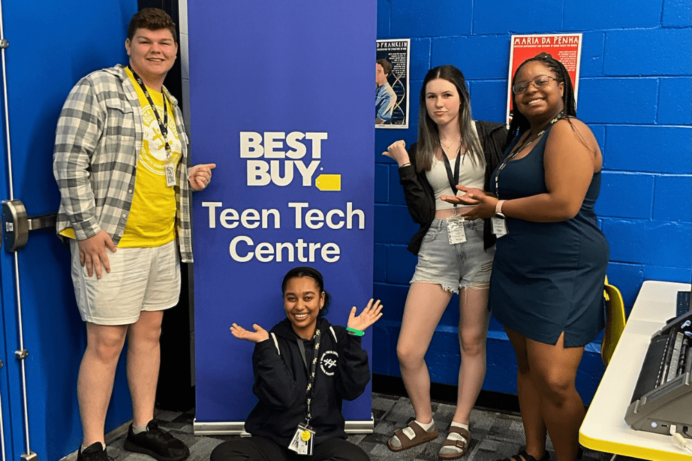 best-buy-teen-tech-centre Four smiling young people posing playfully around a blue "Best Buy Teen Tech Centre" banner in a room with blue walls.