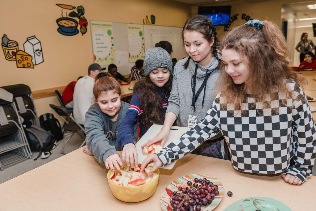 Four children and a staff member gather around a table, reaching for sliced apples and grapes in a BGC Club kitchen