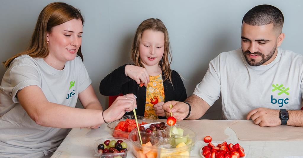 A child and two BGC volunteers sitting at a table, smiling while making fruit skewers with strawberries, grapes, and melons.
