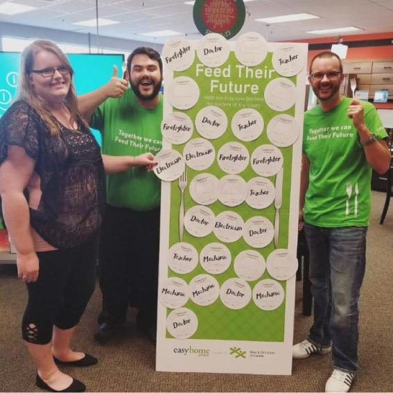 Three smiling staff members pose next to a "Feed Their Future" campaign display board covered in paper plates listing future careers.