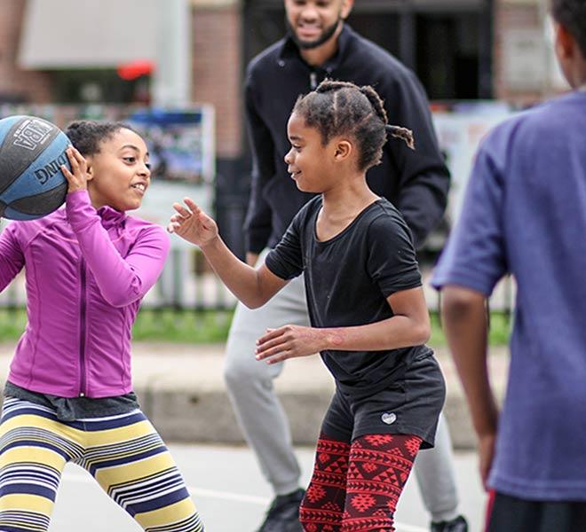 Two young girls playing basketball outdoors with an adult watching in the background.