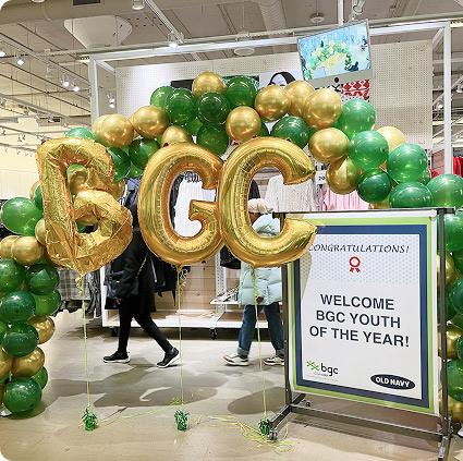 A festive display in a store featuring a large arch of green and gold balloons, with large gold foil balloons spelling "BGC" and a sign that reads "CONGRATULATIONS! WELCOME BGC YOUTH OF THE YEAR!