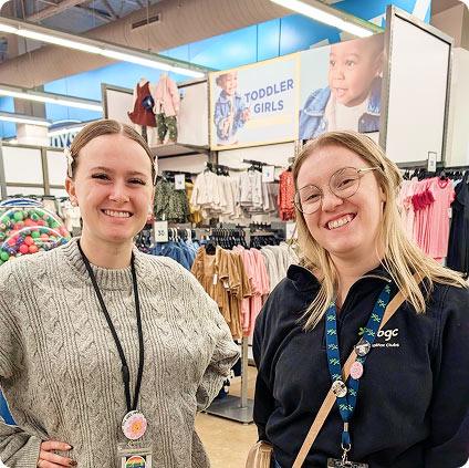 Two smiling young women wearing lanyards pose together in the toddler clothing aisle of a retail store.