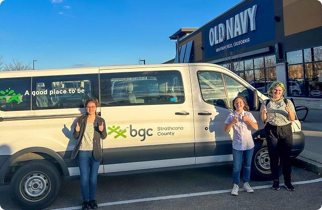 Three young people giving thumbs up in front of a white BGC Strathcona County van parked outside an Old Navy store.