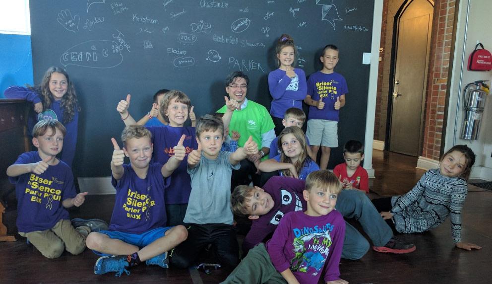 Group of children in purple shirts and an adult in a green shirt giving thumbs up in front of a chalkboard wall.