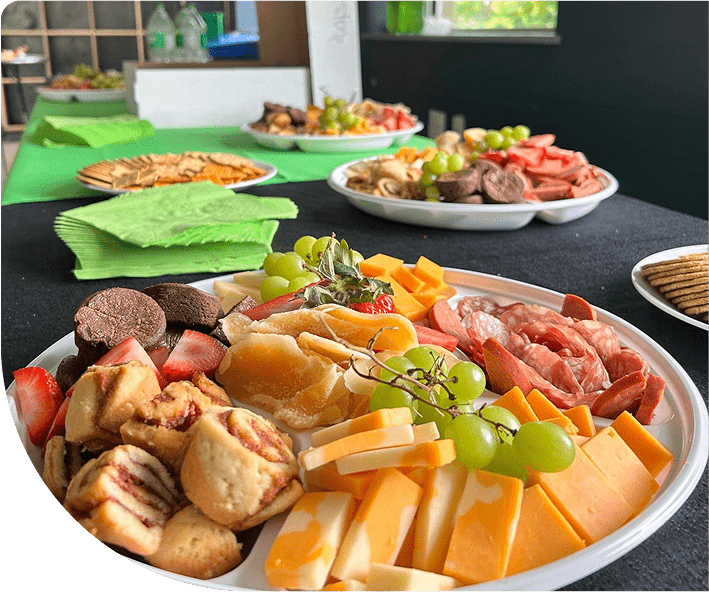Buffet table set with platters featuring assorted cheeses, meats, fruit, and pastries, next to stacks of green napkins.