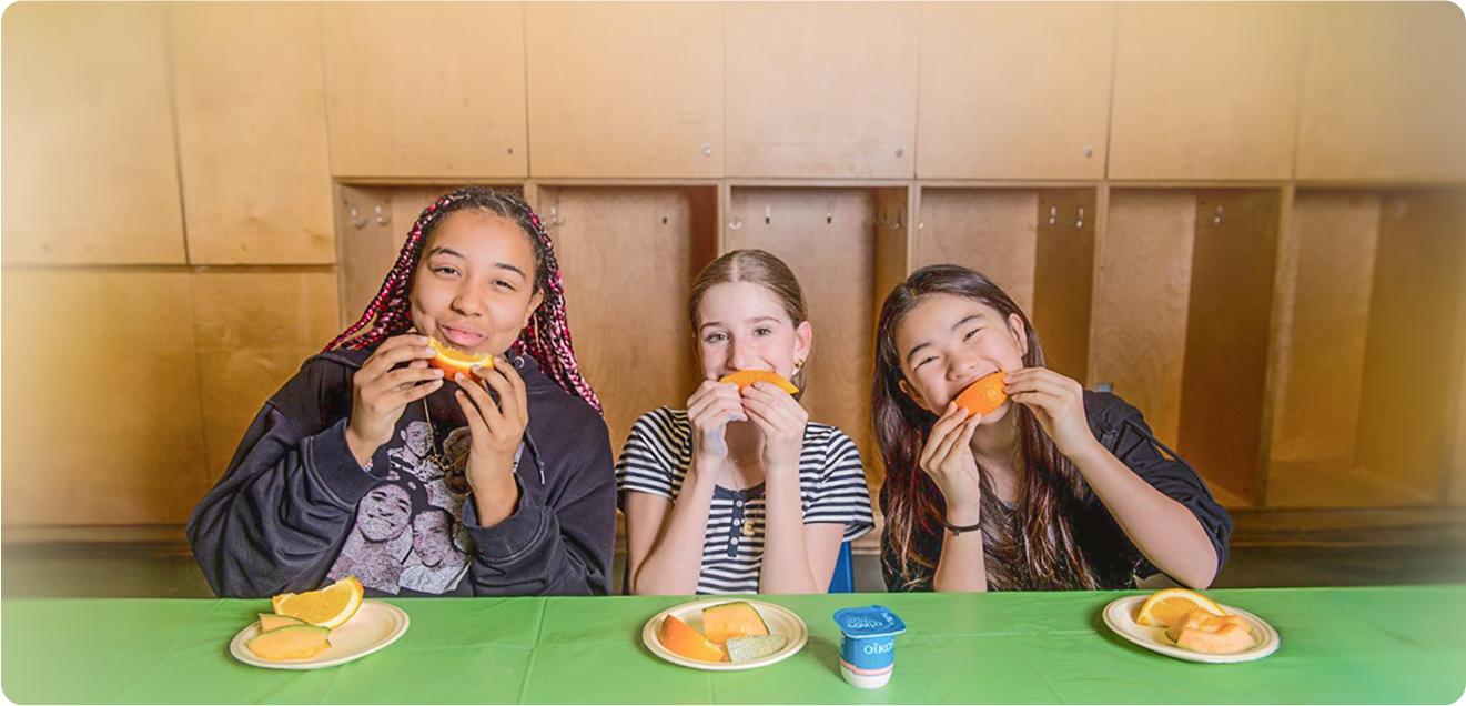 Three smiling girls eating orange slices at a table with fruit and yogurt.