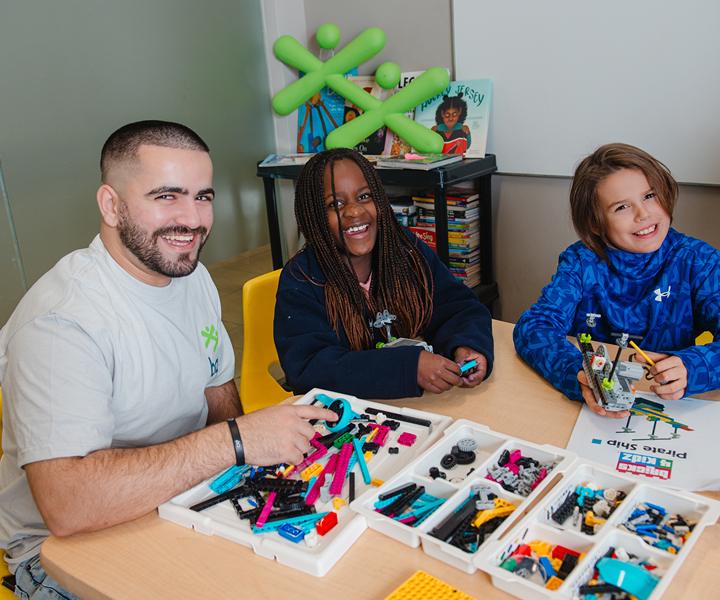 A smiling man and two children work together at a table with colorful plastic building bricks in front of a green BGC logo.