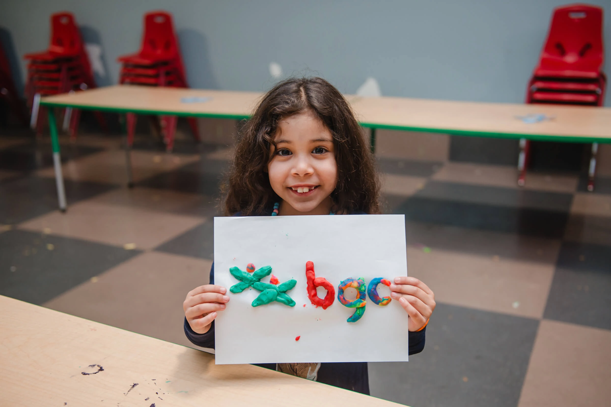 A smiling young girl holds up a handmade BGC logo made of colourful clay letters in a classroom setting.