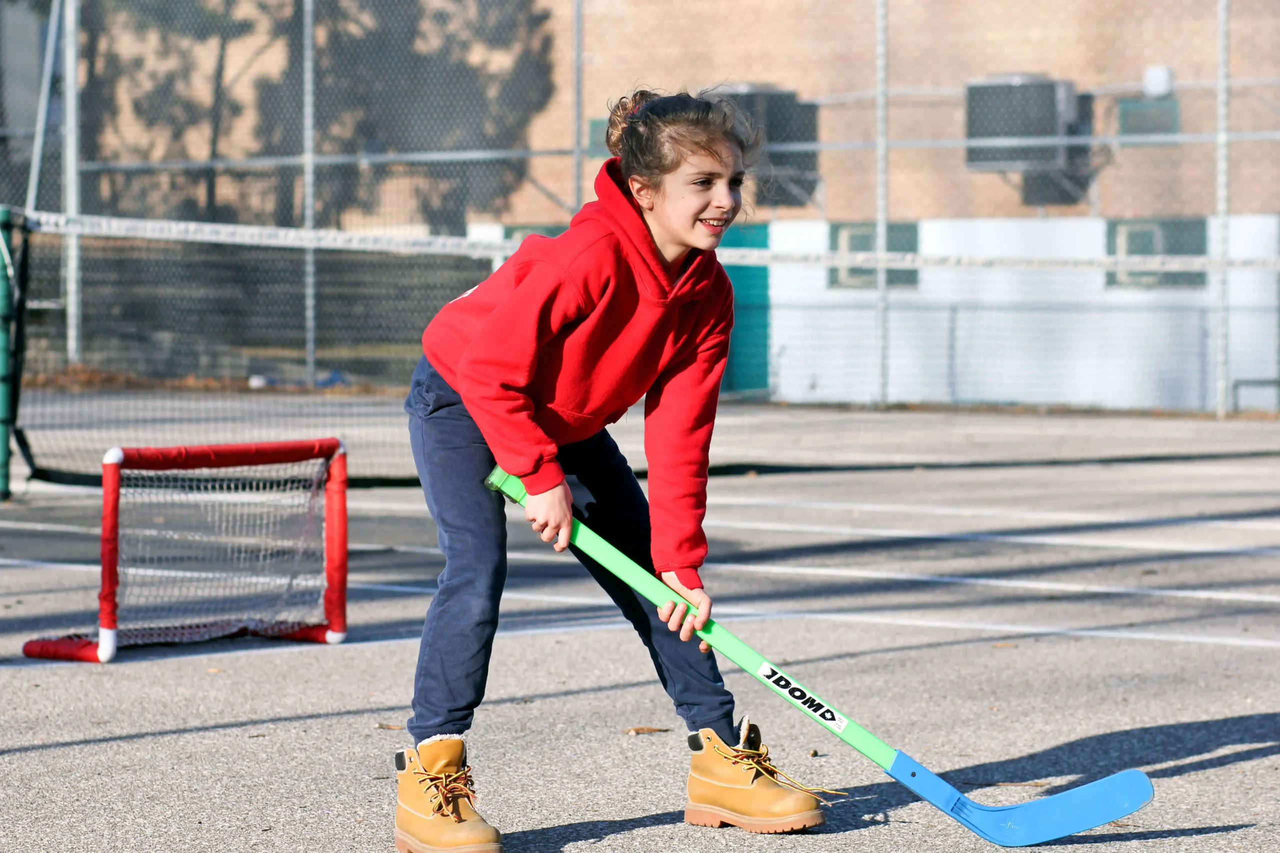 A smiling young girl wearing a red hoodie and boots playing street hockey on a schoolyard with a green and blue stick
