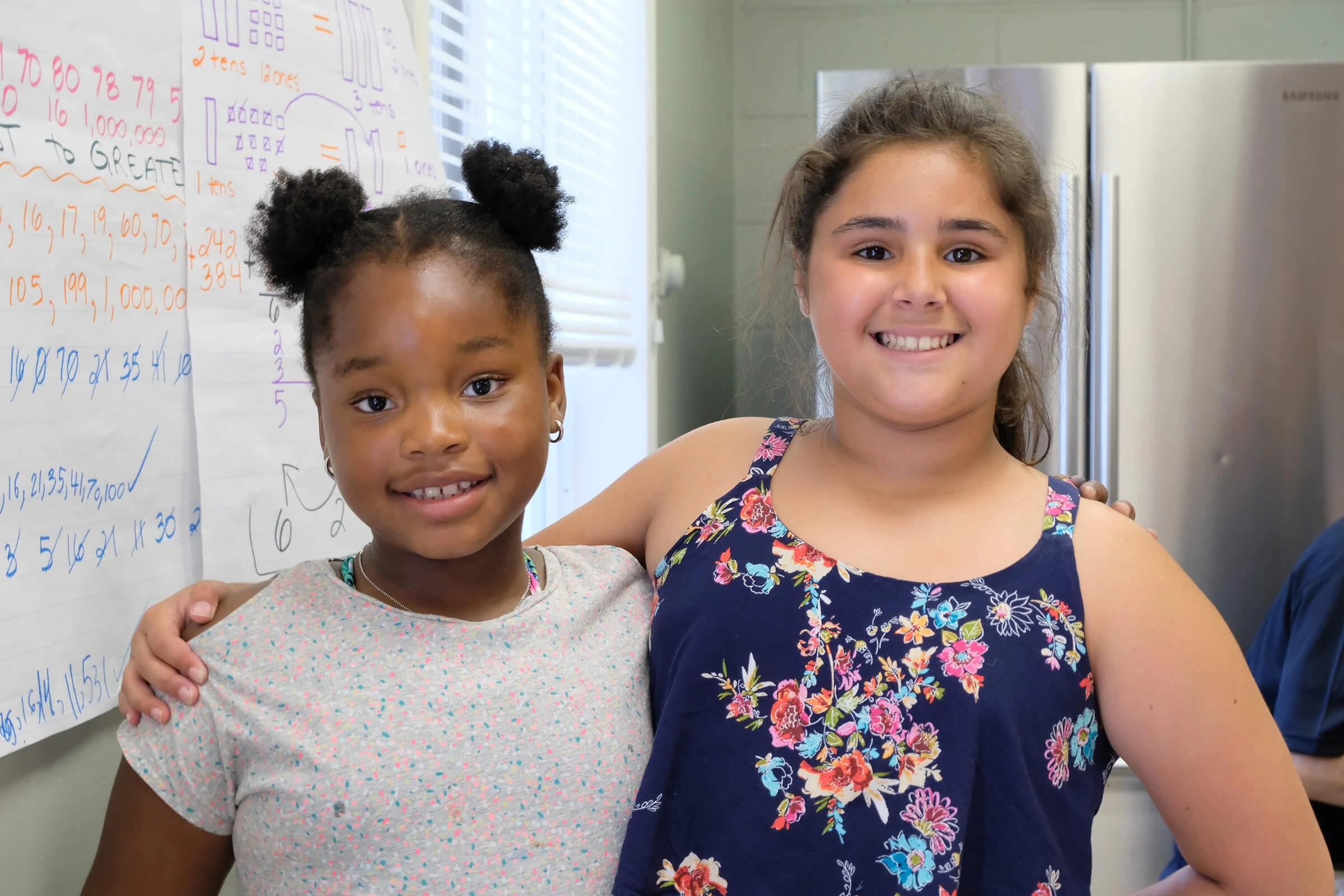 Two smiling girls stand arm in arm in a classroom with math posters behind them, representing friendship, learning, and joy at BGC Clubs.
