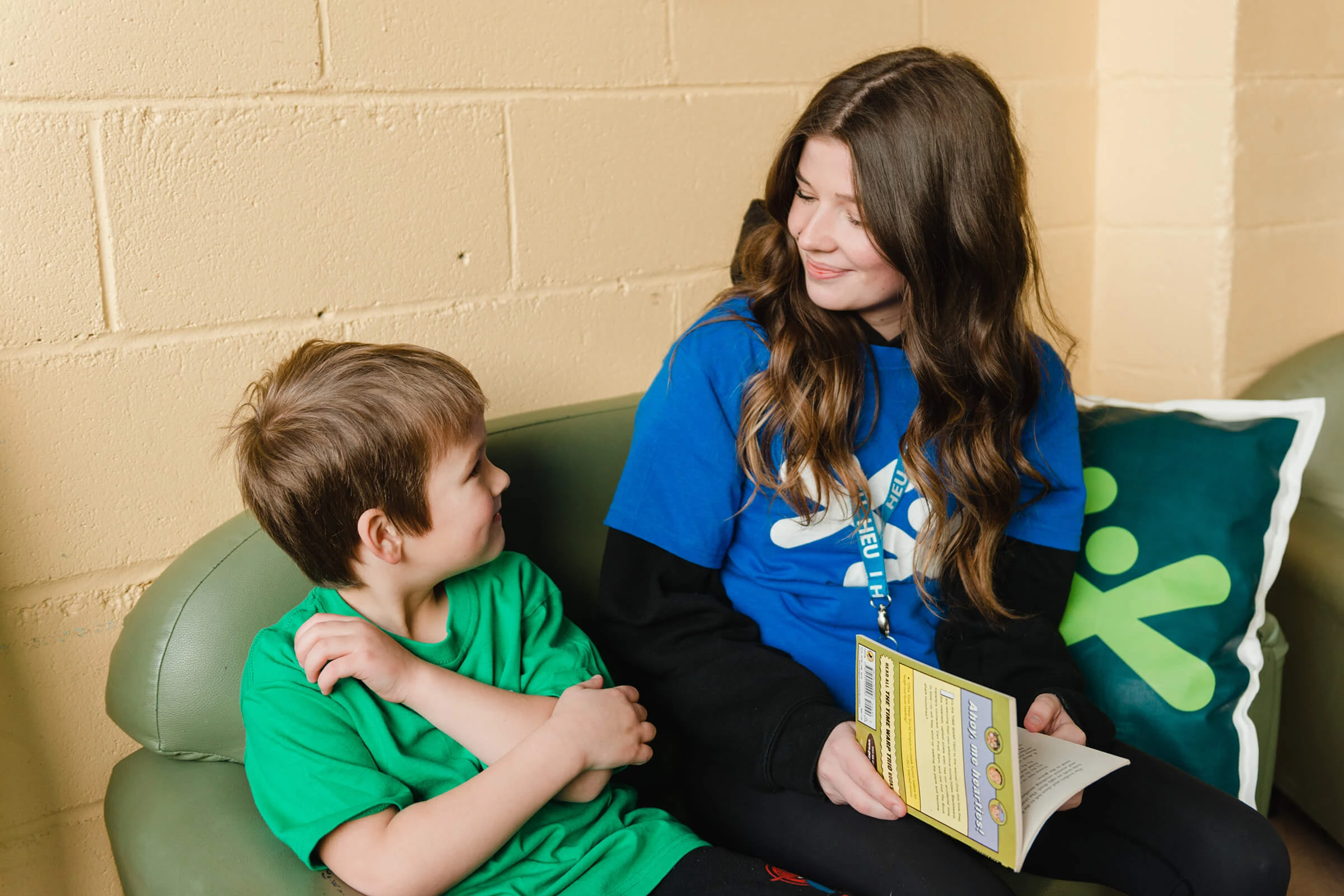 A BGC staff member sits on a couch talking warmly with a young boy, holding a club activity booklet.