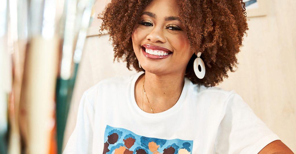 A close-up of a smiling young woman with curly hair, wearing a white t-shirt and large white hoop earrings.