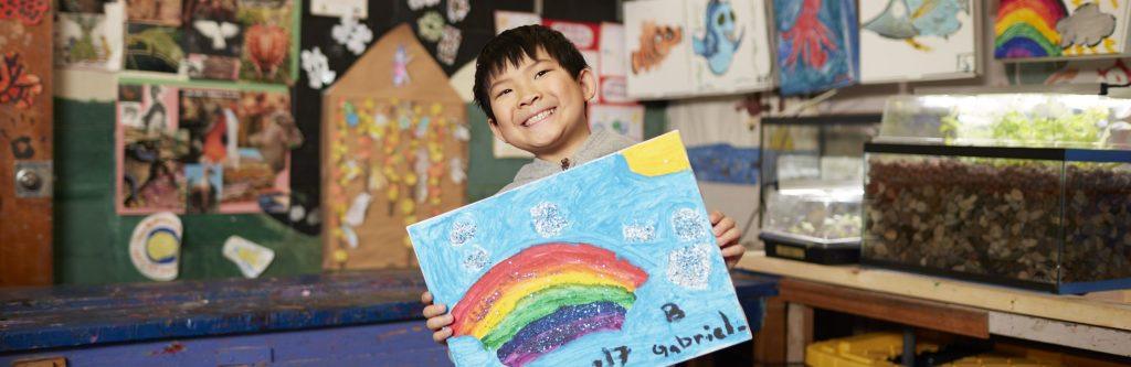 A smiling young boy holds up a colorful painting of a rainbow and clouds decorated with glitter in a classroom filled with art projects.