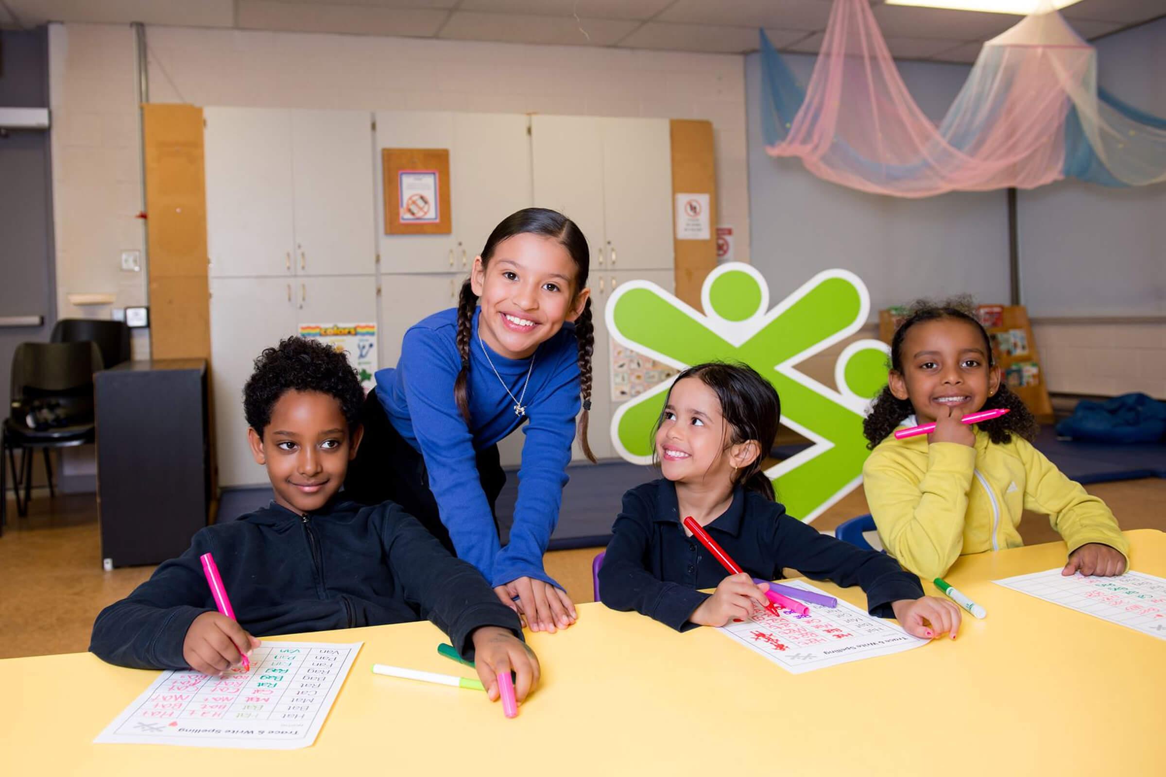 Four children smiling and working on educational worksheets at a BGC Club classroom, with a youth leader standing behind them.