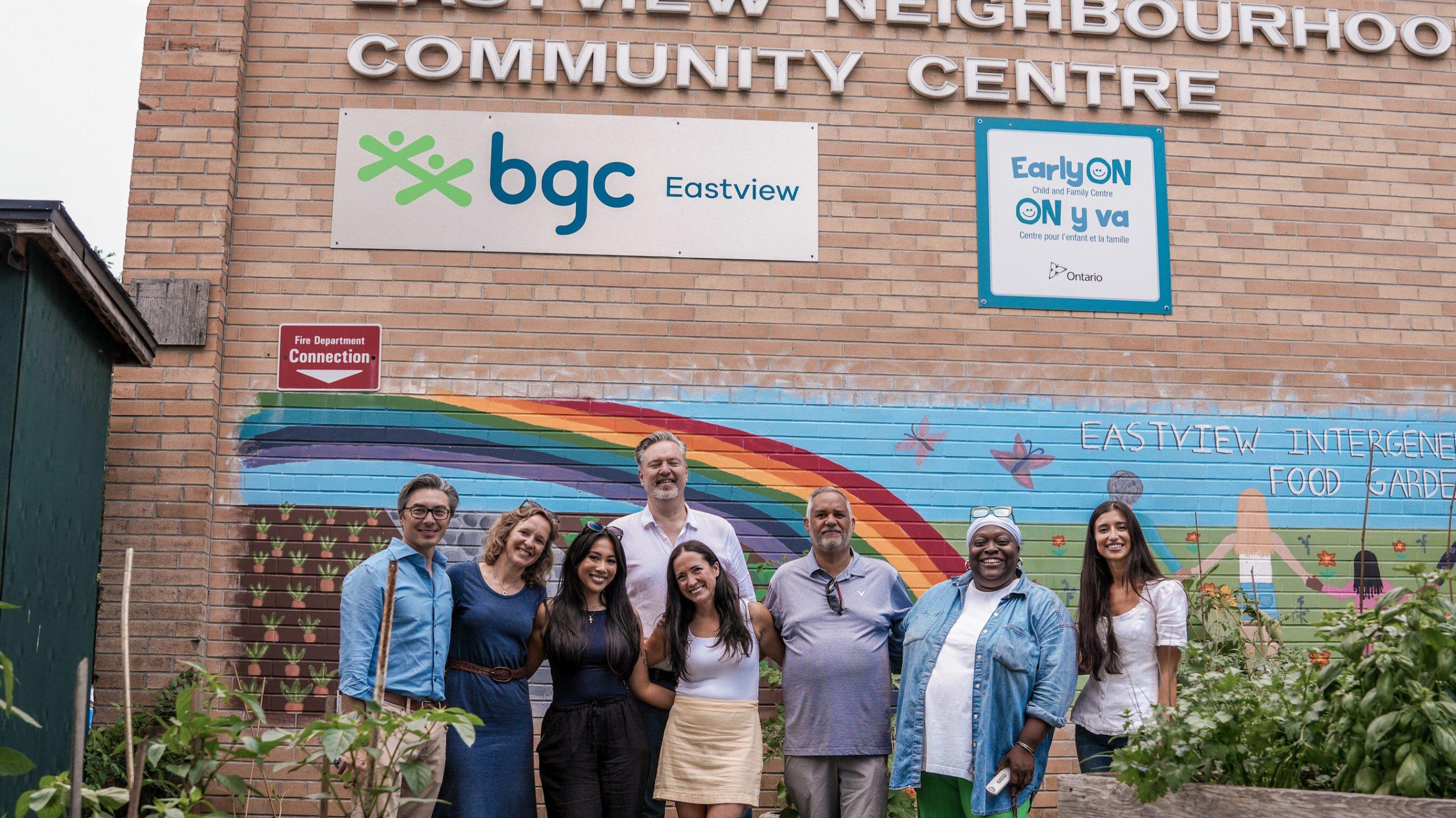 A group of seven diverse adults stands together in front of a brick community center building featuring a BGC Eastview sign and a colorful rainbow mural.