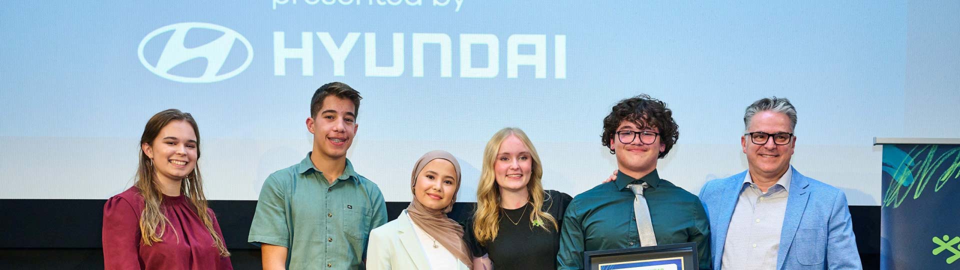 A group of six people, including youth and adults, stand together on a stage in front of a large screen displaying the Hyundai logo.
