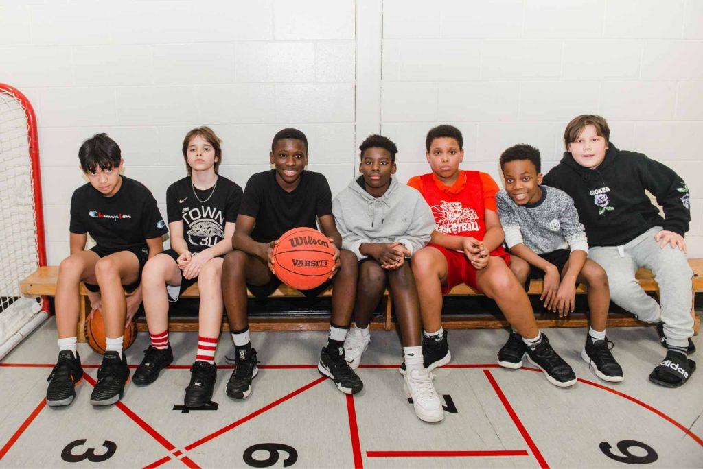 Group of seven young boys sitting on a gym bench, with one holding a basketball.