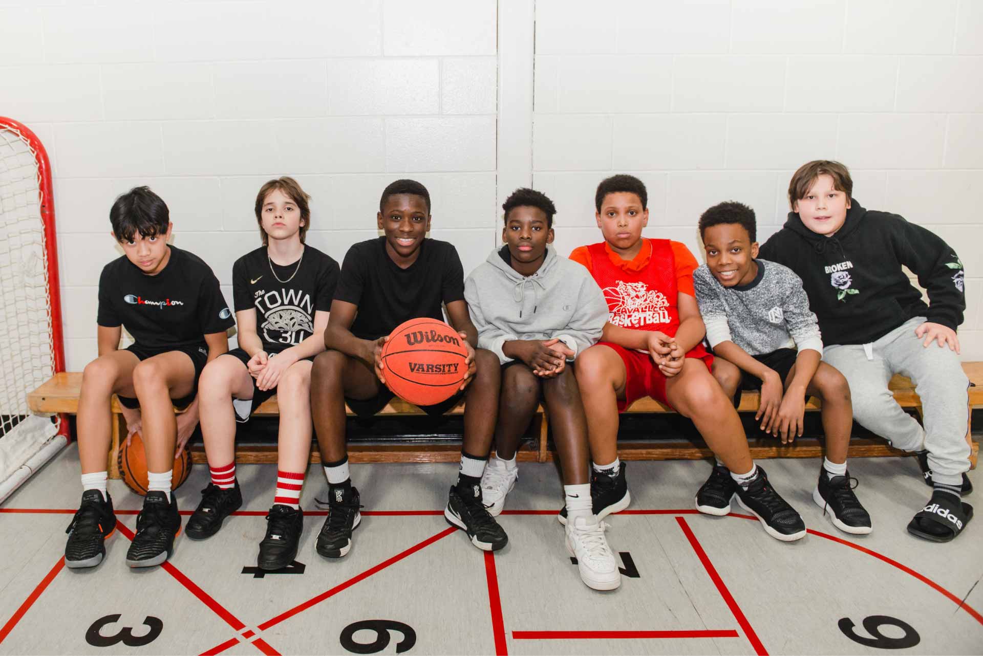 Group of seven young boys sitting on a gym bench, with one holding a basketball.