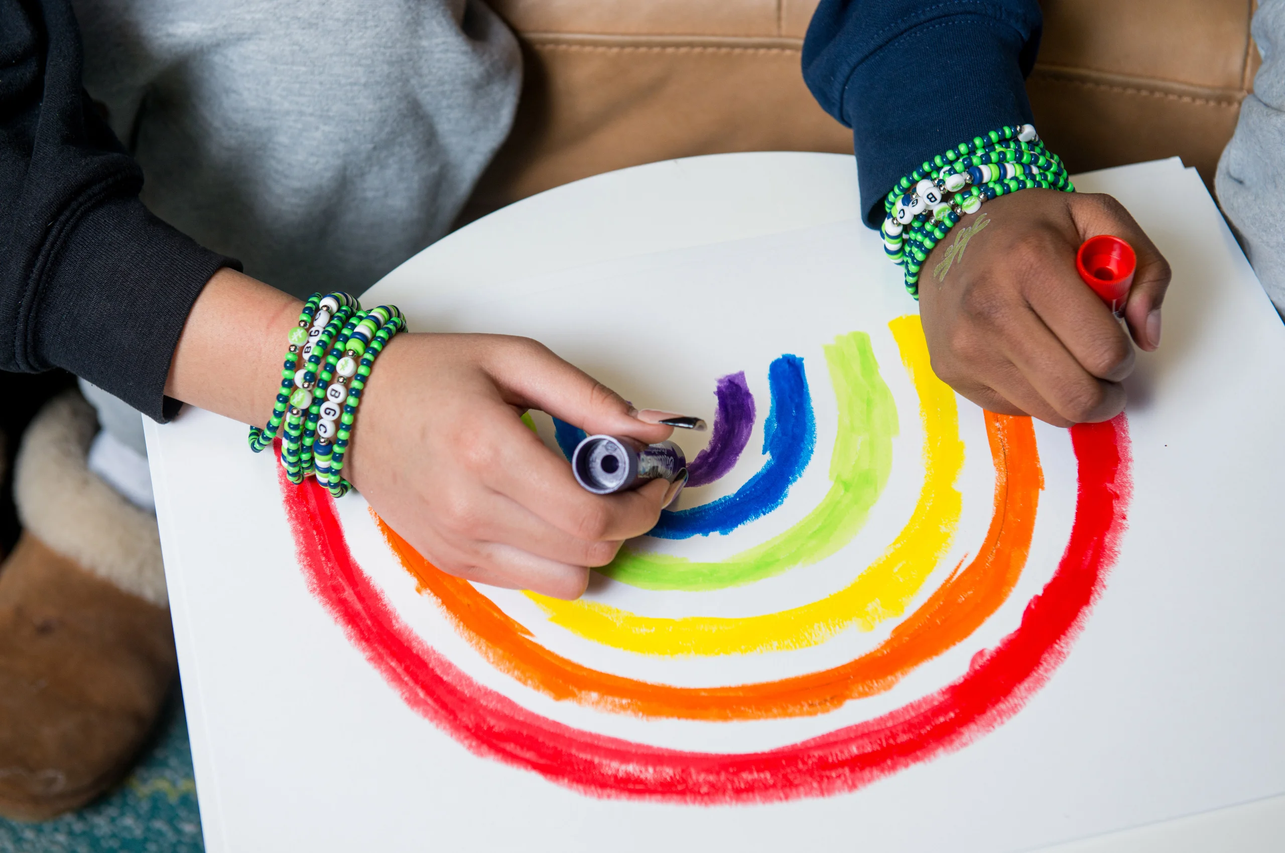 Close-up of hands wearing beaded bracelets drawing a colorful rainbow on paper.