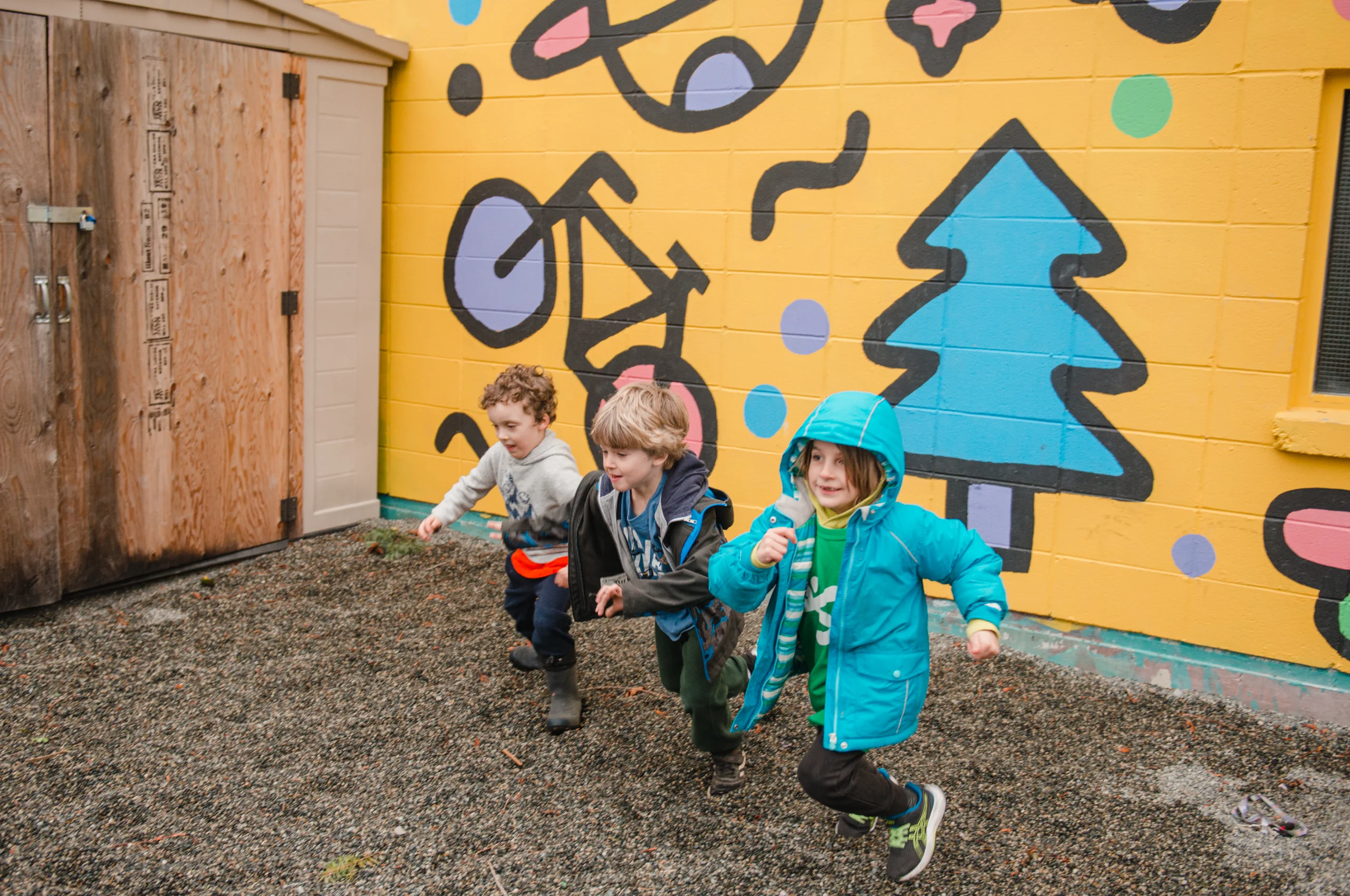 Three children running outside in front of a yellow wall with colorful painted murals.