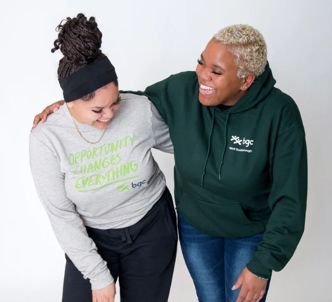 Two women laugh together while wearing BGC apparel, with the woman on the left in a grey sweatshirt reading "OPPORTUNITY CHANGES EVERYTHING" and the woman on the right in a dark green "bgc West Scarborough" hoodie.