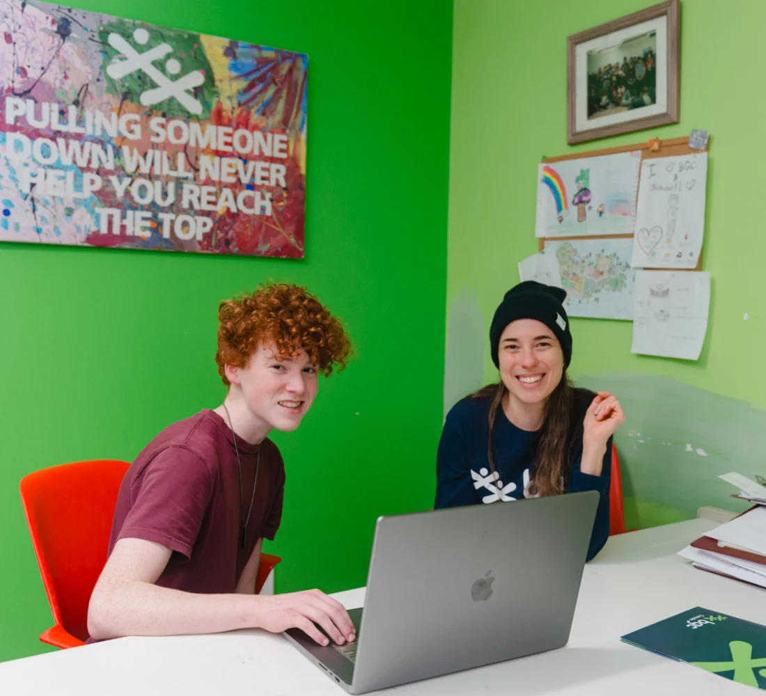A young man and a woman smile while working together on a laptop in a bright green room decorated with youth artwork and an inspirational poster.