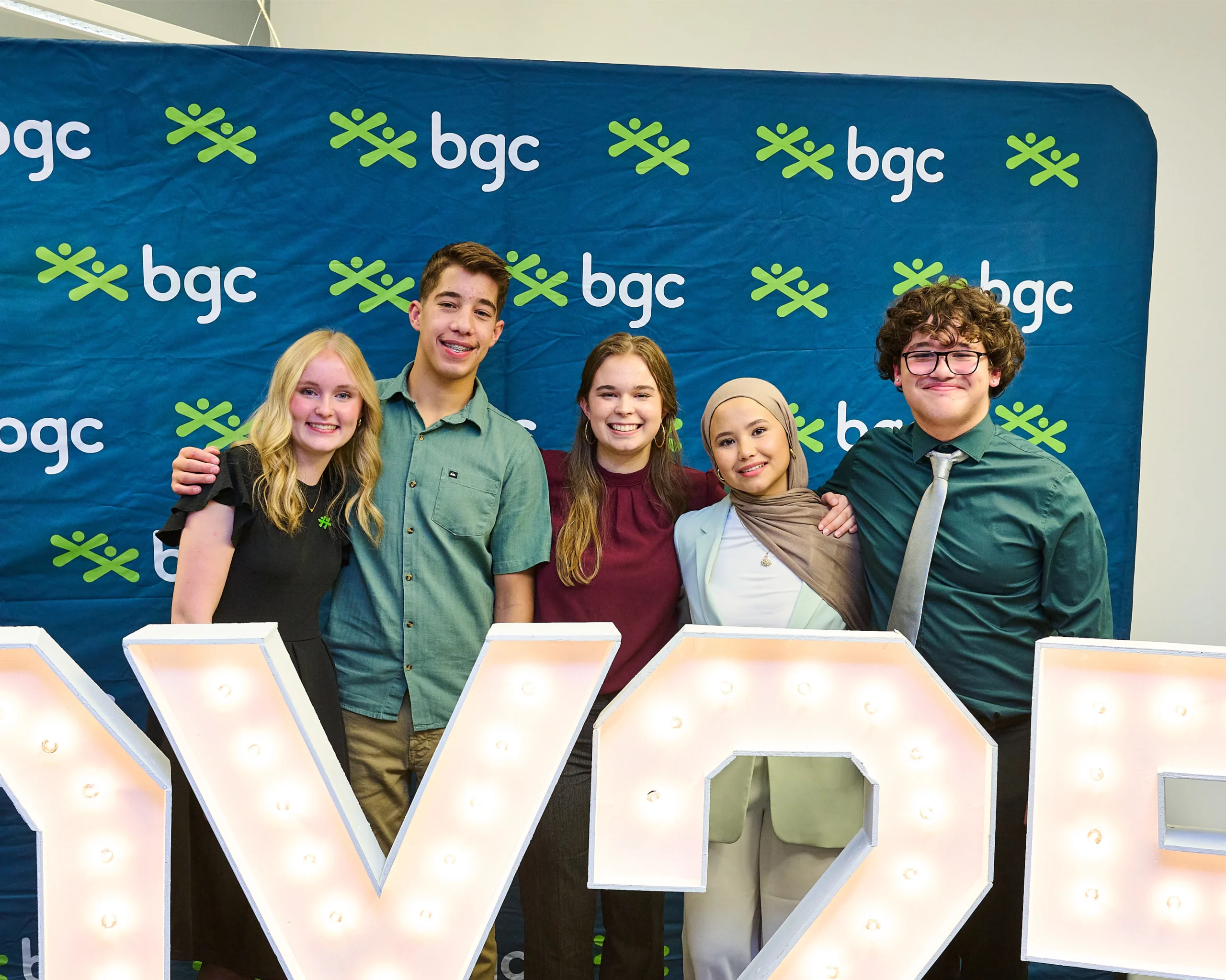 Five smiling young adults pose together with their arms around each other in front of a blue BGC branded backdrop.