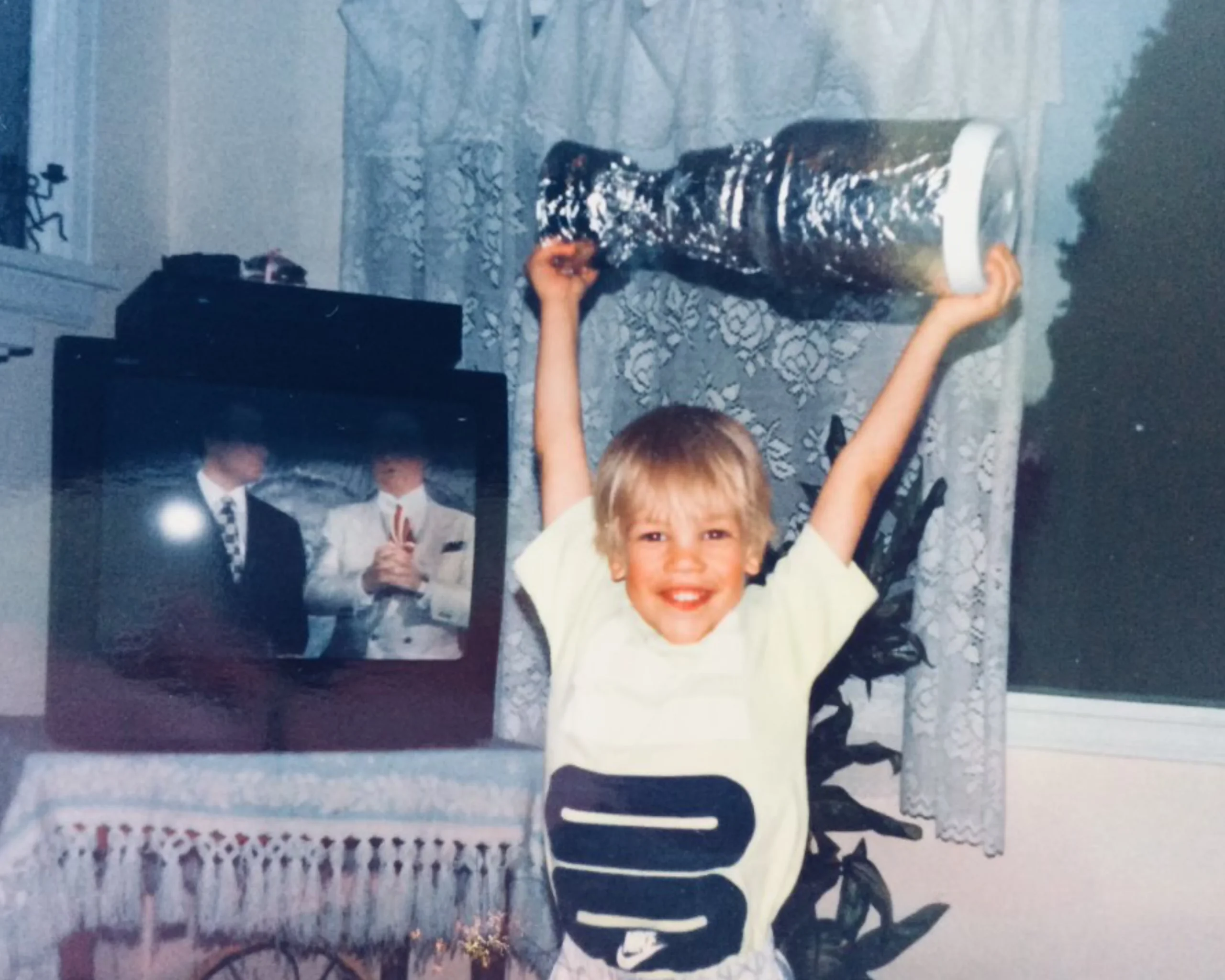 A smiling young boy triumphantly lifts a homemade tinfoil Stanley Cup trophy above his head.