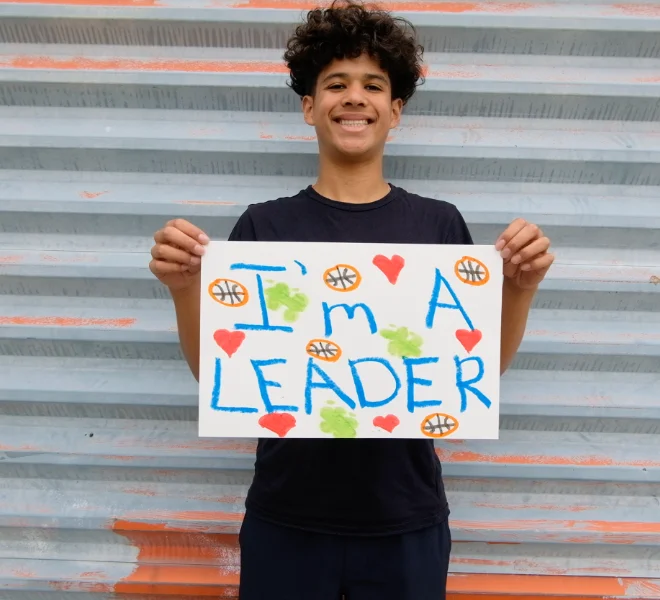 A smiling young man holds a handmade sign that reads "I'm A LEADER" decorated with basketballs and hearts.