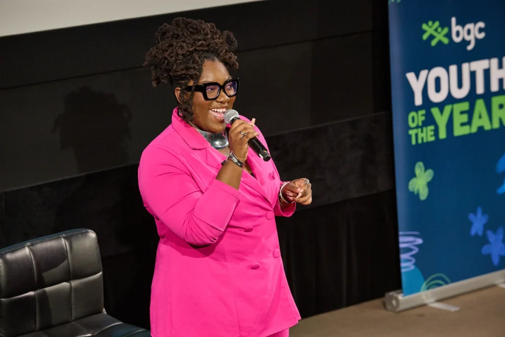 A speaker holding a microphone on stage during a youth leadership event, standing beside a ‘Youth of the Year’ banner.