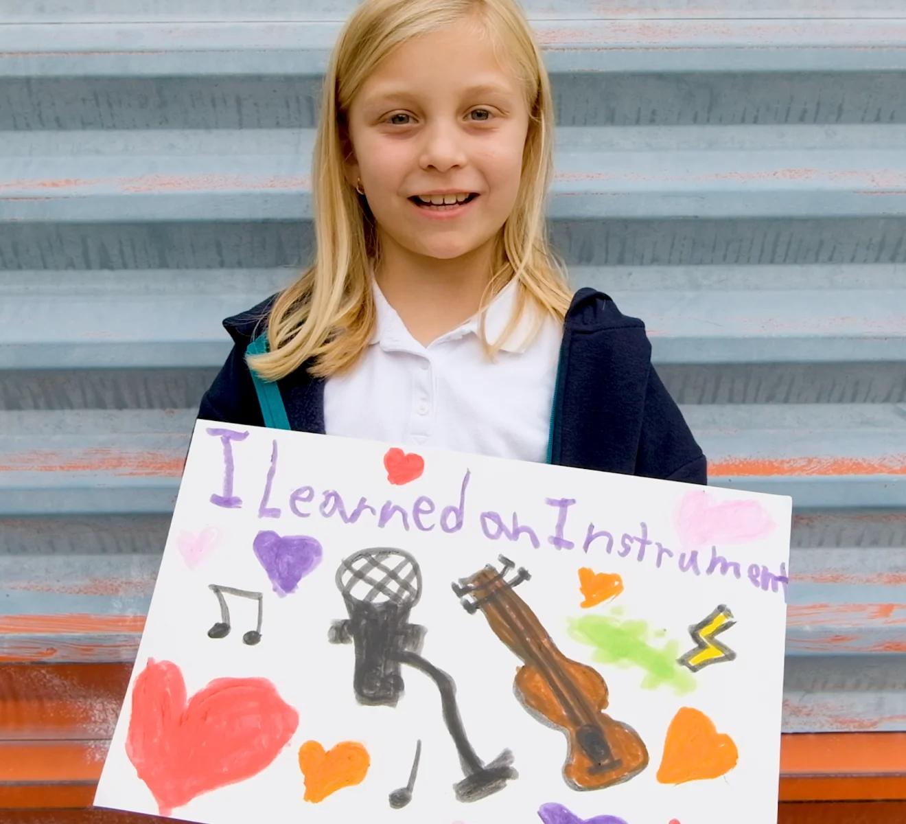 A child smiling and holding a handmade sign that reads ‘I learned an instrument,’ decorated with drawings of music symbols.