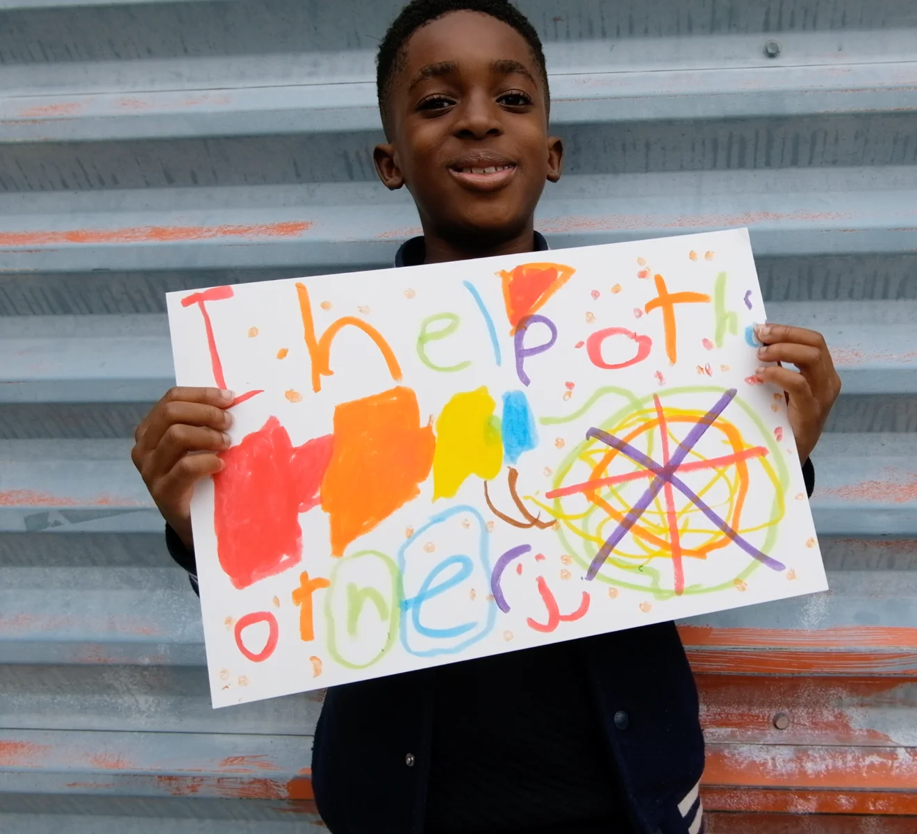 A child smiling and holding a handmade sign that reads ‘I help others,’ decorated with colorful drawings.