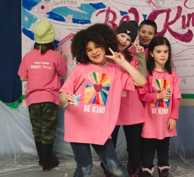 A group of smiling children wearing matching pink "Be Kind" t-shirts pose in front of a mural they are painting.