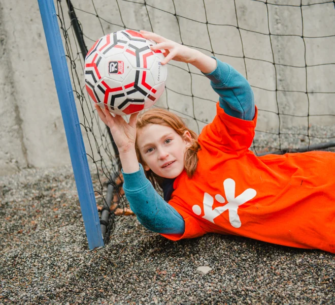 A child playing goalkeeper, reaching up to catch a soccer ball in front of a net.