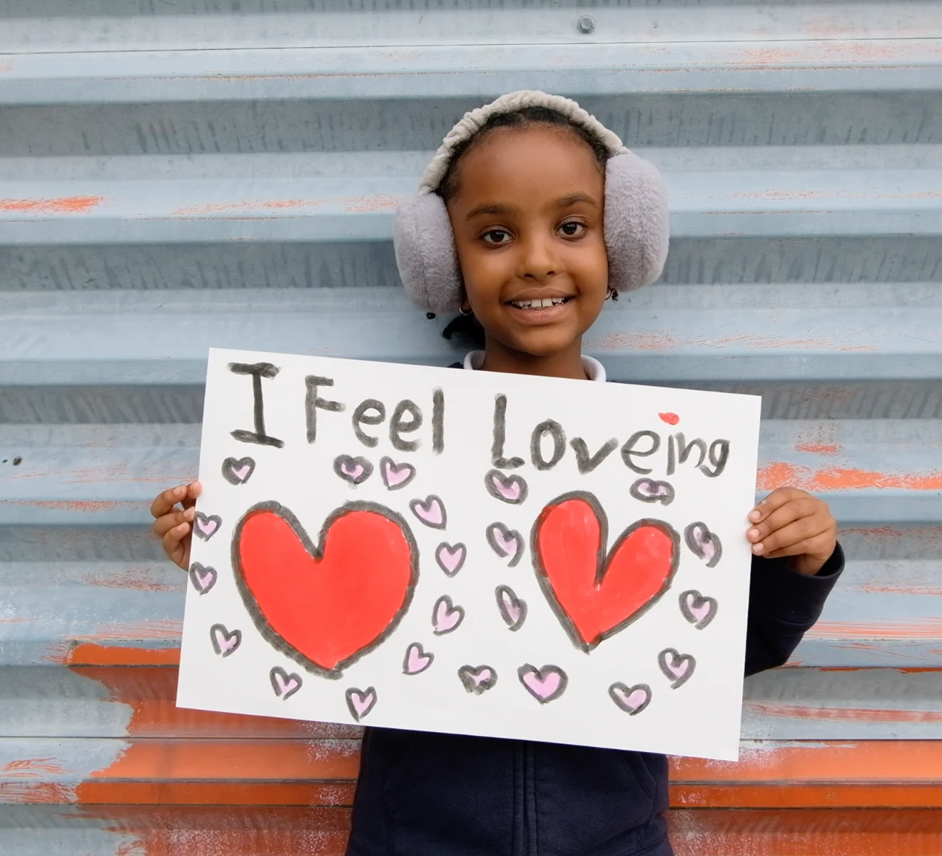 A child smiling and holding a handmade sign that reads ‘I feel loving,’ decorated with hearts.