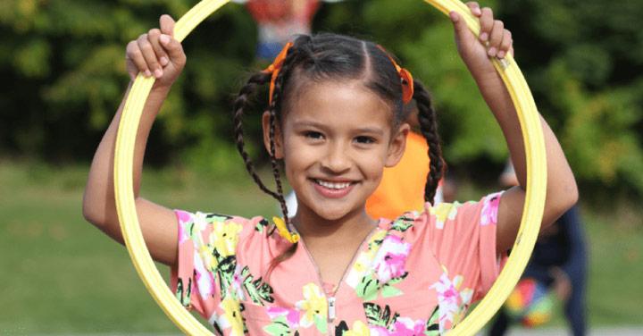 A smiling young girl with braided hair holds a yellow hula hoop around her head.